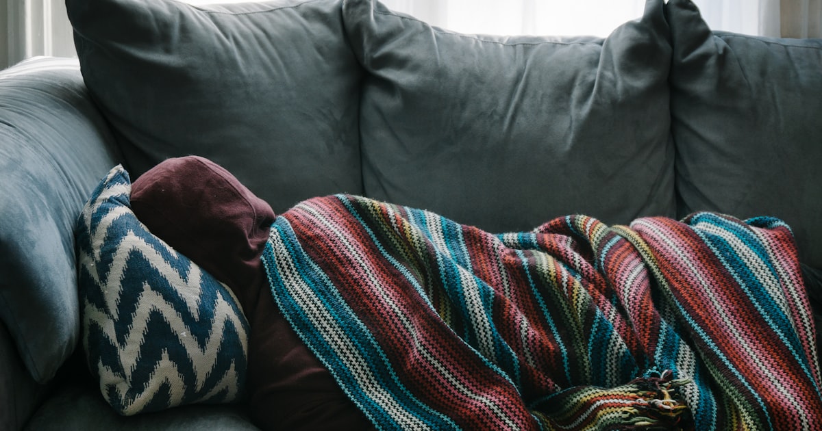 A person resting under a blanket on a gray sofa